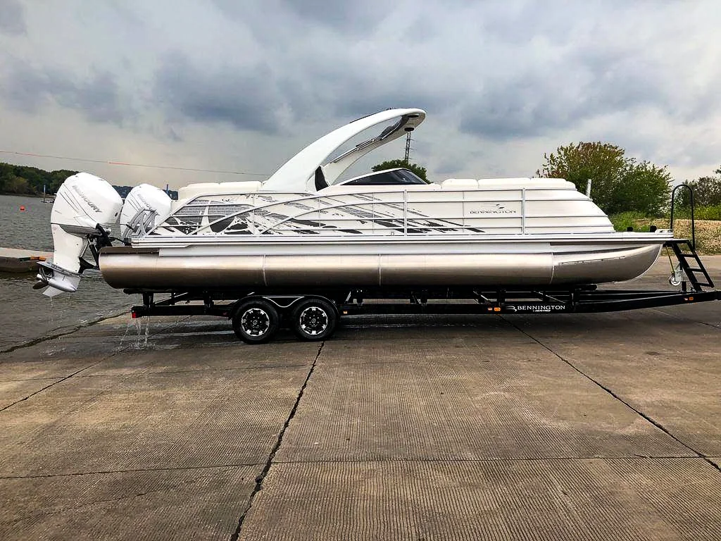 White Bennington tritoon with twin outboards on its trailer at the ramp, the kind of wide load that needs permits to move